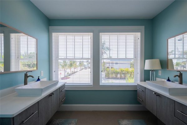 Symmetrical bathroom with twin white vessel sinks, gray wood vanities, and teal walls under bright windows with blinds.