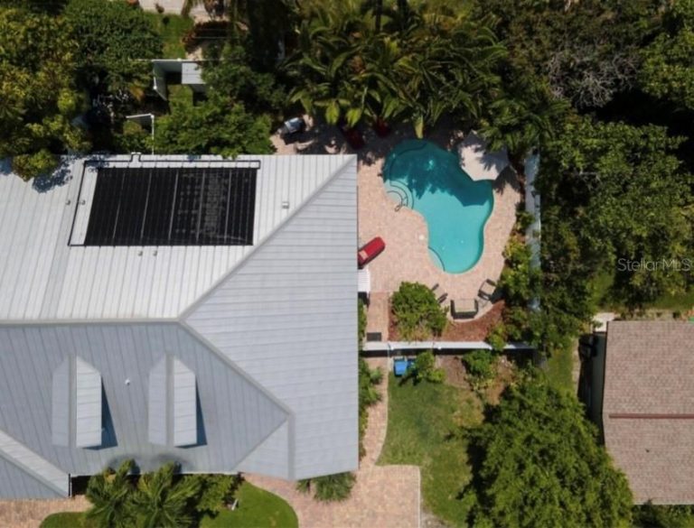 Aerial view of a light-gray roofed house with a kidney-shaped pool in the backyard surrounded by trees and a patio area.