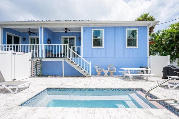 Blue house with white trim and a raised deck overlooking a rectangular pool on a paved patio area.