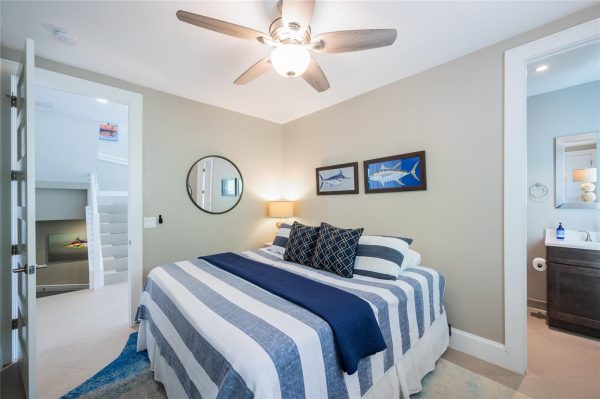 Bedroom with a striped blue-gray bedspread, round wall mirror, two fish pictures, and an adjacent bathroom visible to the right under a ceiling fan light.