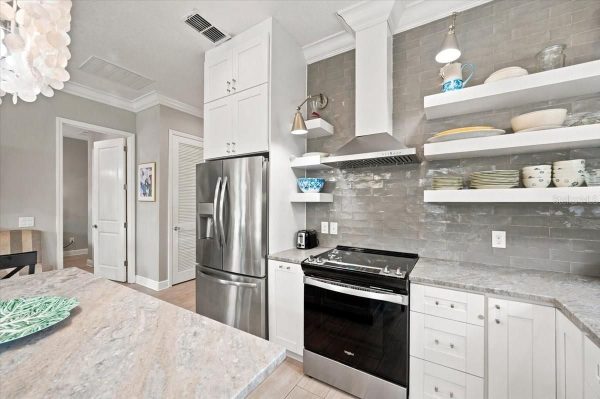 Modern kitchen with stainless steel French-door fridge, gas range, and white cabinets; open shelves with dishes against a gray tile backsplash.