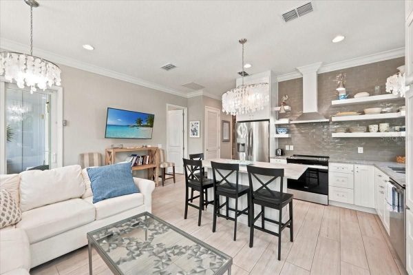 Open-concept living room and kitchen with a white sofa, blue throw pillow, glass coffee table, and a large island with four black chairs under pendant chandeliers.