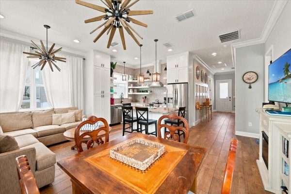 Open-concept living room and kitchen with white cabinets, stainless appliances, and a wooden dining table in the foreground.