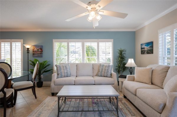 Bright living room with beige sofas, blue accent wall, large windows with white shutters, and a glass coffee table in front of the sofa.
