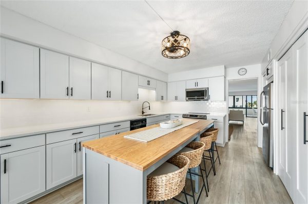 Bright white kitchen with shaker cabinets, black hardware, a wooden island with wicker stools, and stainless steel appliances.