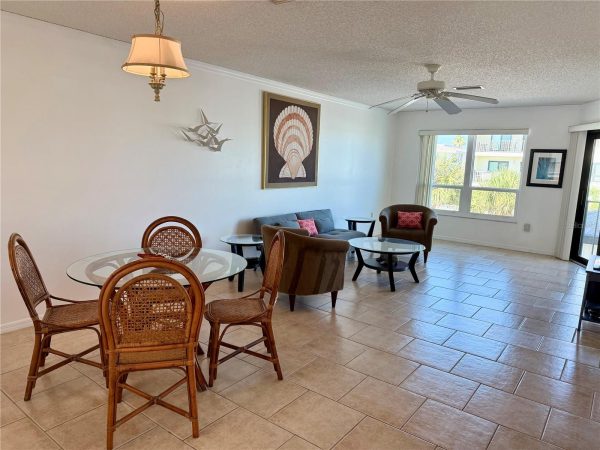 Open-plan living and dining area with a glass round dining table surrounded by four wicker chairs, beige tiled floor, and a seating group of a gray sofa and brown armchairs under wall art and a ceiling fan.