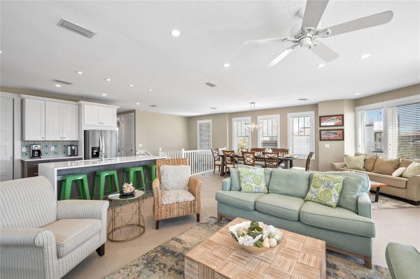 Open-concept living area with white kitchen cabinets, stainless steel fridge, and green bar stools at a long island beside a cozy seating area.