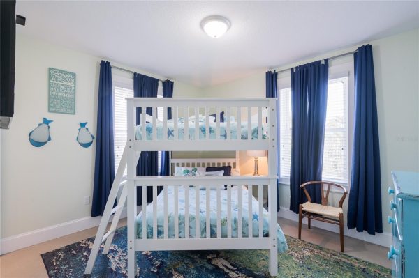 Bright coastal children's bedroom featuring a white bunk bed with a ladder, sea-themed bedding, navy curtains by two windows, a small wooden chair, and a colorful area rug.