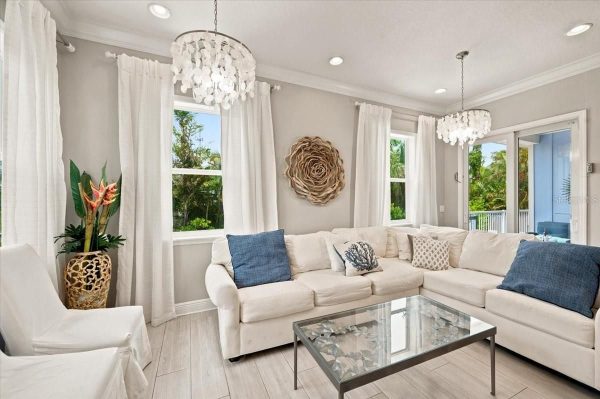 Bright living room with a white sectional, blue pillows, glass coffee table, chandeliers, and large windows with white drapes.