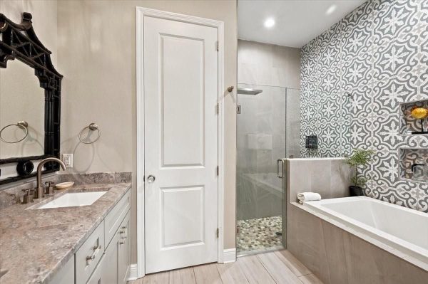 Bathroom with a gray vanity and granite countertop, brass faucet, and a black ornate mirror; white door, glass shower, and a patterned tile wall.