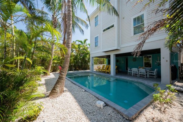 Backyard pool area beside a modern white house with a covered patio and tropical plants.