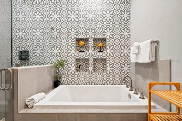 Modern bathroom with a white rectangular tub, patterned tile wall, built-in niches, and a wooden bench beside it.