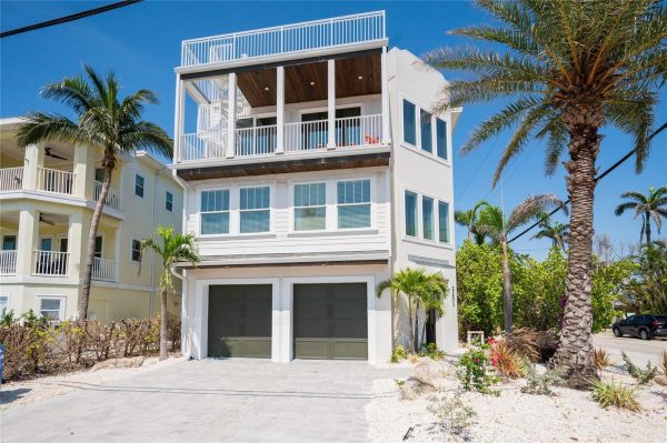 Coastal three-story white house with a large open balcony on the top floor and two gray garage doors on the ground level, surrounded by palm trees under a blue sky.