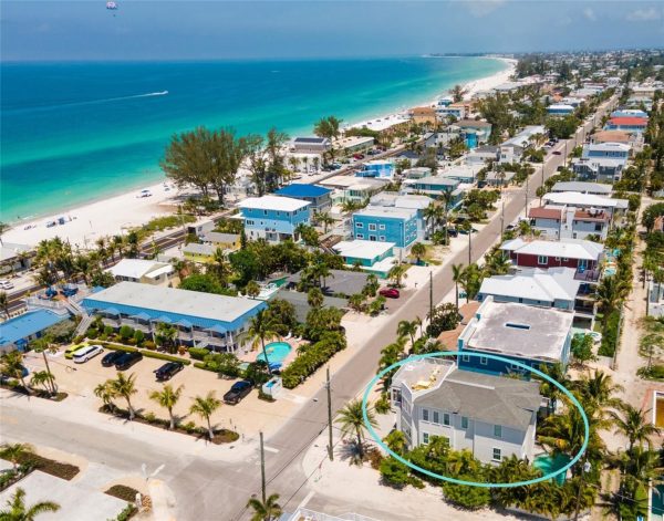 Aerial view of a coastal neighborhood with pastel houses along a palm-lined street and a white-sand beach; turquoise sea in the background, with one house circled in blue in the foreground.
