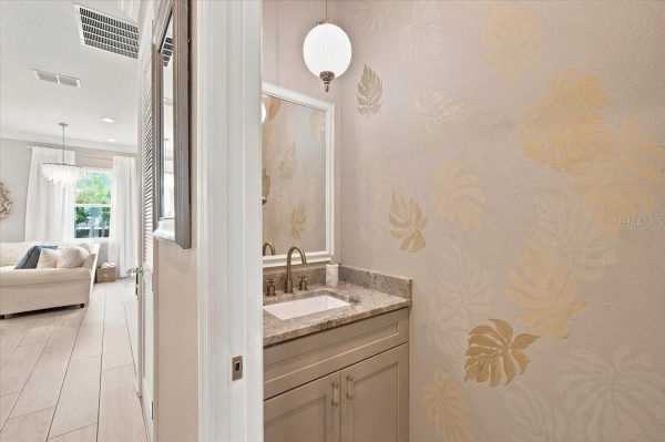 Bathroom vanity with beige marble countertop, brass faucet, and framed mirror next to a gold-floral wallpapered wall, opening to a bright living area with a white sofa.