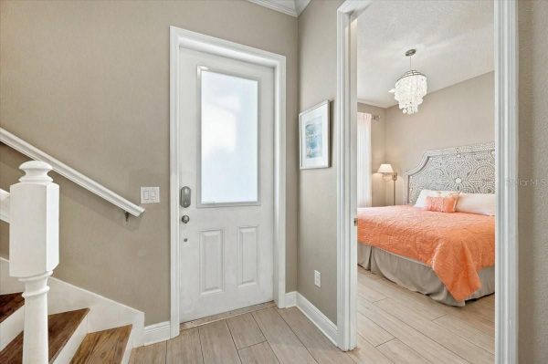 Entryway with a white front door, staircase on the left, and a doorway to a bedroom with a coral bedspread.