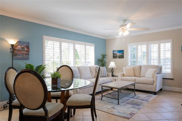 Bright living room with a blue accent wall, beige sofa set, glass dining table, and large white-shutter windows.