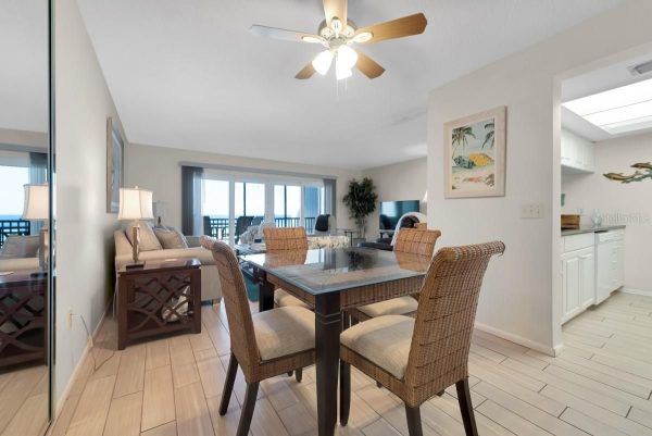 Open-plan dining area with a glass-top table and wicker chairs, beige tile floor, and a living room with a TV in the background near sliding balcony doors.