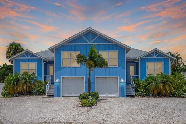 Blue coastal-style house with white trim, two garage doors, and symmetrical peaked roof, with palm trees and a gravel driveway at sunset in the background