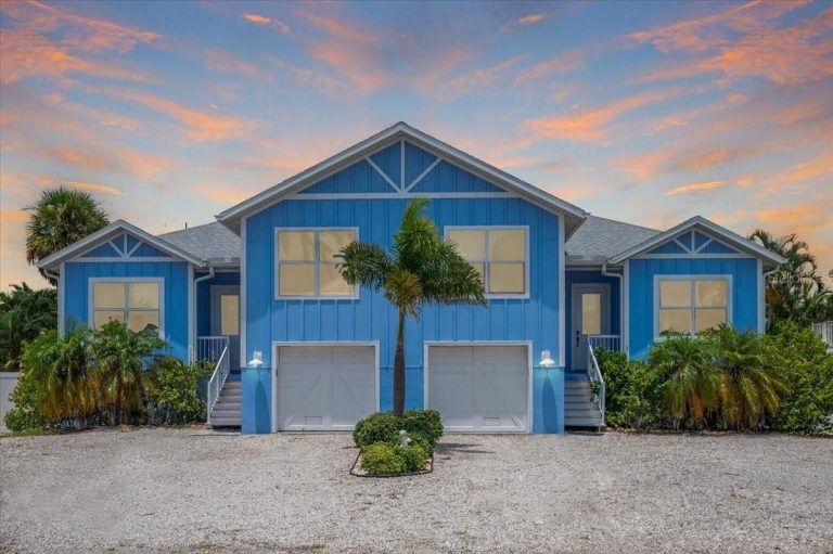 Blue coastal-style house with white trim, two garage doors, and symmetrical peaked roof, with palm trees and a gravel driveway at sunset in the background