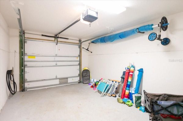Garage interior with a white sectional door, stored items along the right: folding chairs, beach umbrellas, and pool toys. A blue duct runs along the ceiling and a hose reel is mounted on the left wall.