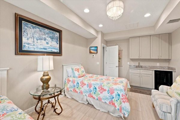 Bright hotel-style twin bedroom with coral reef–patterned bedspreads, a glass-topped side table, and a gold lamp between two single beds. A kitchenette area with cabinets and sink is visible in the corner.