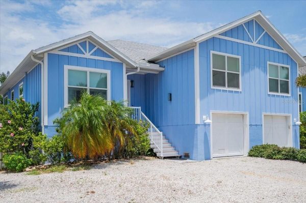 Two-story blue house with white trim, front stairs, and two white garage doors, tropical shrubs in the yard.