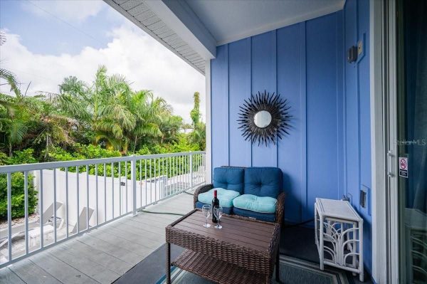 Blue-walled balcony with wicker sofa, teal cushions, and a glass-tinished coffee table holding a wine bottle and two glasses, tropical garden beyond.