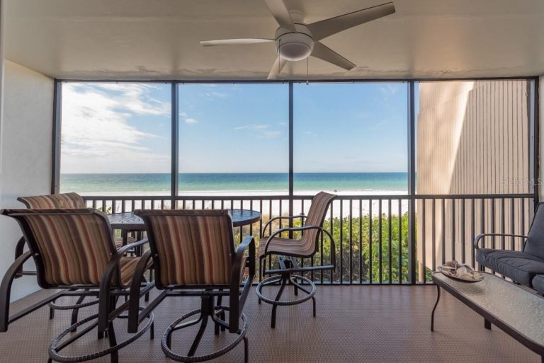 Balcony seating with striped chairs around a table, overlooking a sandy beach and blue ocean beyond a glass railing.