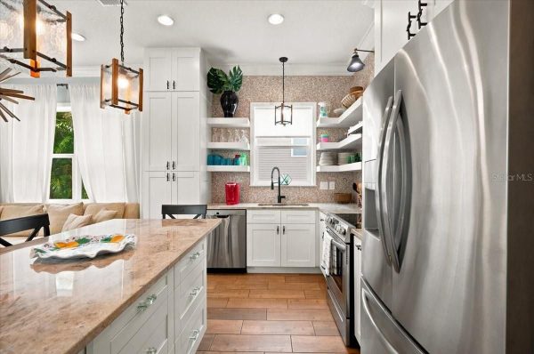 Bright modern kitchen with white cabinets, beige countertop island, stainless appliances, and open shelving near a dining area on the left.