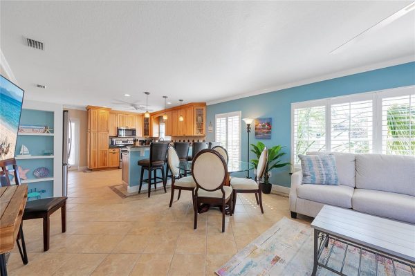 Open-concept living area with light wood kitchen cabinets, island and round glass dining table by a blue accent wall; large windows and a gray sofa create a bright, coastal room.