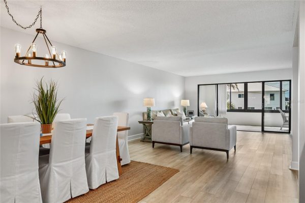 Open living-dining space with a wood dining table, white slipcovered chairs, a potted plant, a gray sofa set, and sliding glass doors to a balcony.