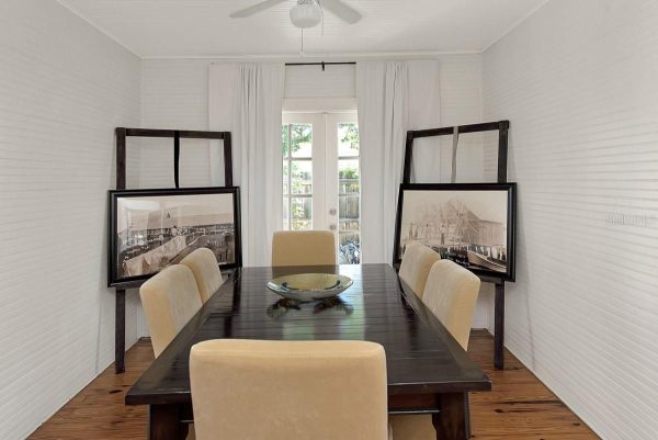Dining room with a long dark wooden table, beige upholstered chairs, white paneled walls, and two large framed photos on easels along the sides, near glass doors at the back.