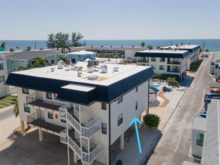 Aerial view of a beachside residential area with white buildings and dark blue roofs; ocean in the background and a marked ground-level entrance under a building.