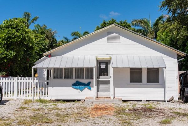 Small white wooden house with a metal awning, front door, and a blue fish decoration on the wall in a tropical setting.