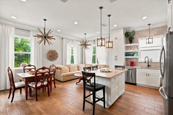 Open-concept kitchen and dining area with a large island, white cabinets, stainless steel appliances, and a beige sectional sofa under bright windows with white curtains.