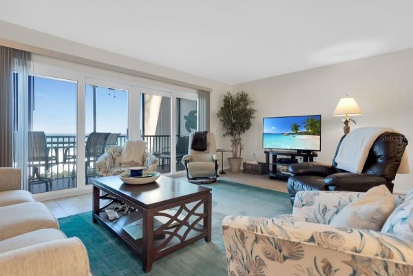 Living room with a teal rug, glass coffee table, and beige seating facing a TV; sliding glass doors open to a balcony with ocean view.",