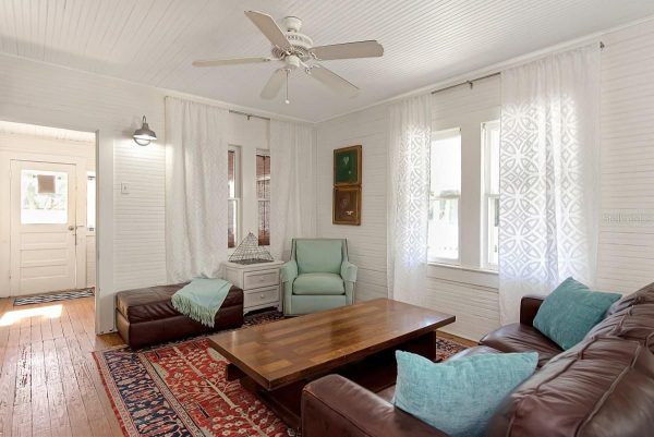 Bright living room with white walls, wooden floor, brown leather sofa, and a wooden coffee table under a red patterned rug.