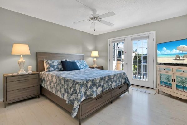 Coastal hotel bedroom with a king bed, blue seashell quilt, two lamps on wood nightstands, and French doors opening to a balcony.