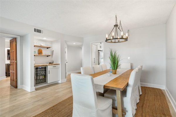 Bright dining room with a long wooden table, white upholstered chairs, and a circular chandelier; a small bar area with shelves is on the left.