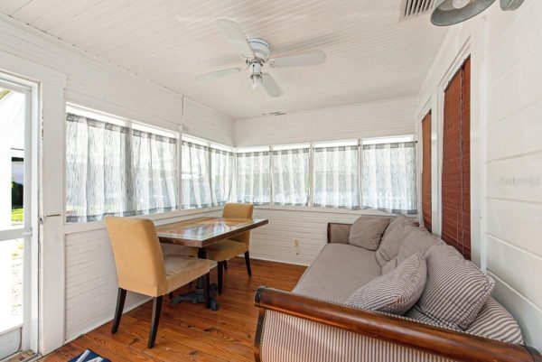 Bright enclosed sunroom with white paneled walls, long windows with sheer curtains, a striped sofa, and a glass-top dining table with beige chairs.