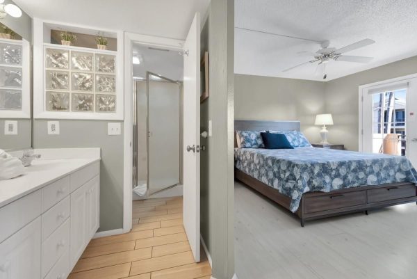 Hotel suite: left shows white bathroom vanity with glass-block window; center doorway to shower; right shows bedroom with blue floral bedding and balcony door.