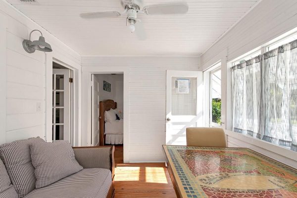 Bright white sunroom with a striped sofa, mosaic-tile table, ceiling fan, and large windows with sheer curtains; doorway opens to a bedroom beyond.