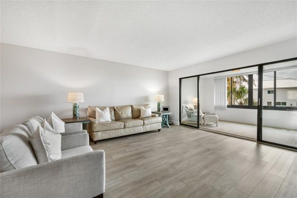 Bright, neutral living room with light gray chairs and a beige sofa, two end tables with lamps, and a sliding glass door to a balcony. Outside are palm trees and a building.